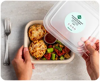 A person opens a meal container holding two cheese-topped muffins, roasted vegetables, and sauce in a cup. The label reads: Cheesy Veggie Melt with Spaghetti Squash Marinara with nutrition details. A fork lies nearby.